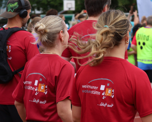 Kurz vor dem Start stehen alle Läufer bereits in Position. Die Läufer stehen mit dem Rücken zur Kamera. Auf den Shirts ist das Logo der Weimarer Wohnstätte zu sehen.